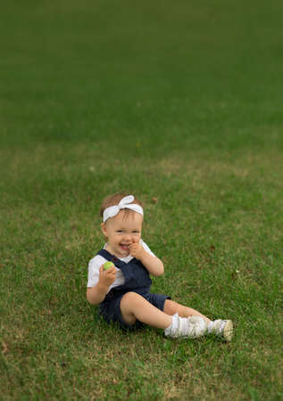 A little girl in a denim jumpsuit sits in the park on the grass and eats an apple. Healthy, complete and proper baby food.の写真素材