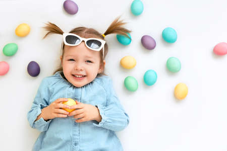 Cute laughing girl on a white Easter background with colored eggs. A child wearing sunglasses.