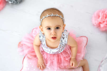 A little girl in a shiny party dress looks up. Cute baby's face close-up. Silver jewelry on the head of a little girl. Expressive big eyes of a girl.の写真素材