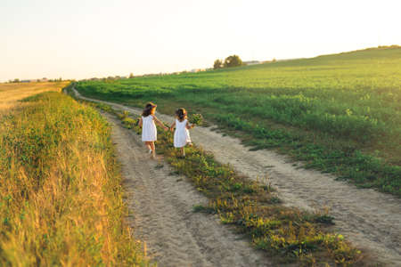 Two girls run along a country road. The road between the wheat and clover fields. Landscape at sunset. Girls in white dresses and with a wreath on their heads.の写真素材