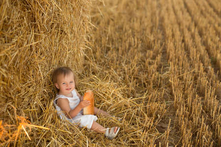A little girl in a field next to a haystack. A hungry child eats bread in a field at sunset. A healthy happy child in harmony with nature.の写真素材