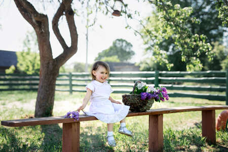 Girl sitting on a bench in the village with a basket of lilacs. The child is happy and cheerfulの写真素材