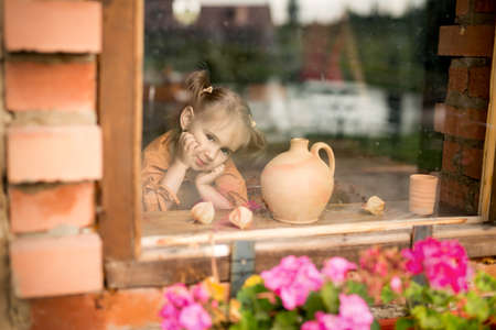 The girl sits alone at home by the window. A child looks out of the window. Dirty, unwashed glass. Dirty, dirty windows.の写真素材