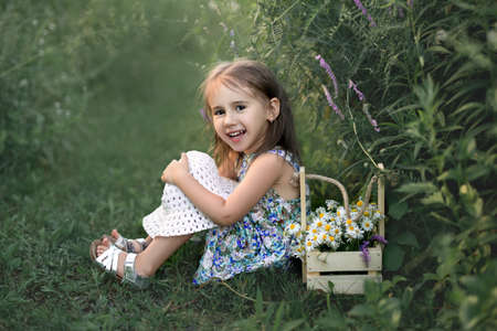 Little girl sits on a green meadow and laughs. The child put the hat on his knees. There is a basket of daisies nearby.の写真素材