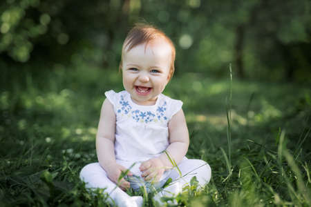 The baby sits on the green grass in the park and laughs. The first two teeth are visible when the baby smiles. Healthy baby laughs. care, caring,の写真素材