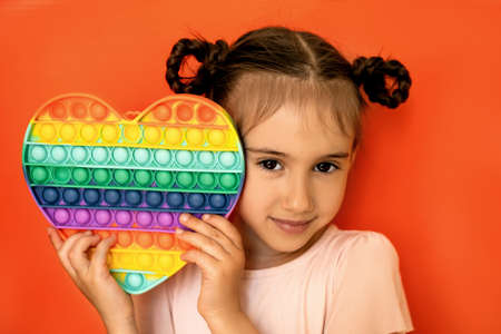 Cute girl with pigtails holding a big heart popit toy. Studio portrait of a child on a red background. Expressive beautiful brown eyesの写真素材