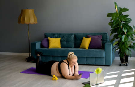 Home fitness classes and Pilates workouts, online classes at home. An overweight woman lazily lies on a sports mat instead of training and plays on her phone. A woman communicates online by phoneの写真素材