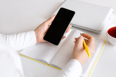 A business woman at her desk with a mobile phone and a pen in her hands. A laptop, cold tea and a clean notebook on the table. Mockupの写真素材