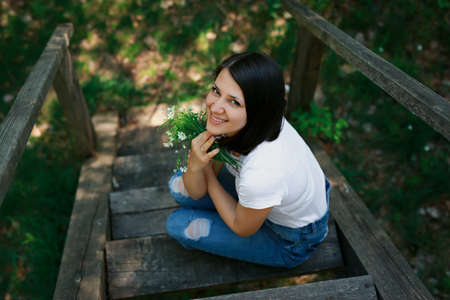 A young smiling woman with a bouquet of white wild flowers is sitting on an old wooden bridge and looking up.の写真素材