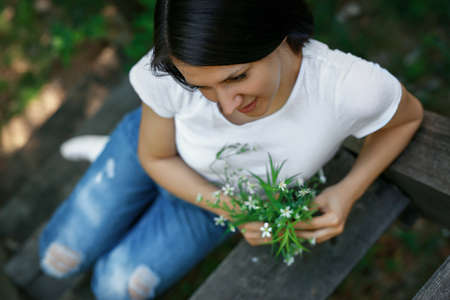 A woman with a bouquet of white wild flowers sits thoughtfully dreaming on an old wooden footbridgeの写真素材