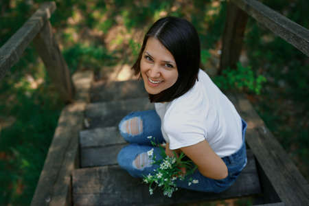 A laughing woman is sitting in the park on the old wooden stairs with a bouquet of white wild flowers.の写真素材