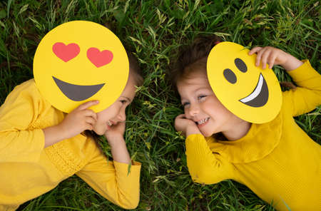 Two funny little girls are lying on the lawn covering part of their faces with emoticons and laughing merrily. World emoji Day. Anthropomorphic smile Face.の写真素材