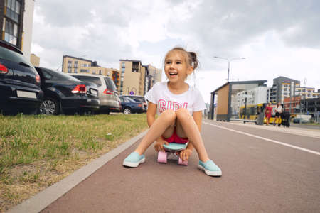 A little girl is sitting on a skateboard on a city street and laughing merrily. The child is learning to skate and so far without success. Entertainment in the city. Active babyの写真素材