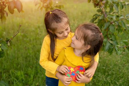Two girls in the park hugging each other holding a heart made of puzzles - a symbol of awareness about autism. Support and love in the family. Equality and friendship.の写真素材