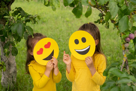 Two children in the park are standing with their faces covered with happy emoticons and showing hand gestures. Funny temperamental girls. Communication.の写真素材