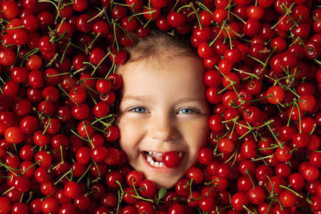 The face of a happy, cheerful child surrounded by a large number of fruits of red, ripe, juicy cherries. The girl holds a cherry in her mouth. Large expressive eyes of a child.の写真素材