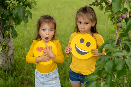 The sisters are holding the faces of happy emoticons in their hands. Two girlfriends in the park under an apple tree look with surprise and perplexity with his mouth openの写真素材