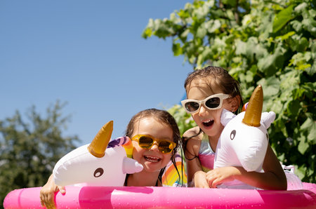 Two little wet girls in sunglasses are having fun in an inflatable pool with a circle in the form of a unicorn. Water splashes. Water activities on a hot summer dayの写真素材