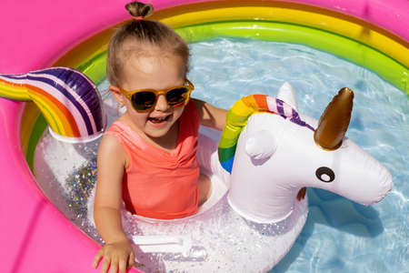 A cute little girl in sunglasses is swimming with a unicorn-shaped lifebuoy in a small inflatable pool. A child splashes in clear clear water on a hot summer dayの写真素材