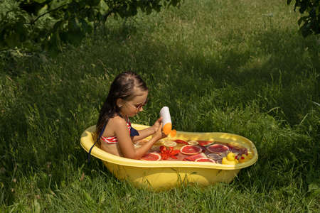 A girl bathes in a bathtub with rubber toy ducklings in nature in the sun and pours shower gel into water with citrus fruits and a lily flower. A product for cleansing and caring for children's skinの写真素材