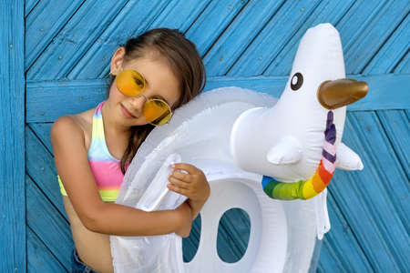 A beautiful girl in yellow sunglasses and a swimsuit is standing by a painted blue wooden board wall with a circle in the shape of a unicorn. Swimming children's circle for the pool or the seaの写真素材