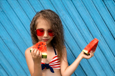 A beautiful girl savors a piece of ripe juicy sweet watermelon with pleasure. Curly stylish girl in red sunglasses on a blue background. Blue wooden plank background. One person. with my eyes closedの写真素材