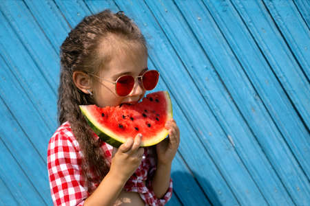 National Watermelon Day. A girl with gathered curly hair and bright red glasses takes a bite of a watermelon. Food on a hot summer day. High sugar content.の写真素材