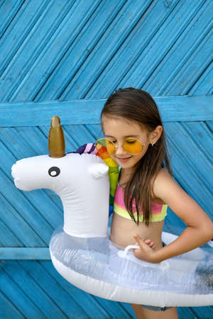A cute beautiful girl in yellow sunglasses and a swimsuit is standing against a blue wooden plank wall with a circle in the shape of a unicorn. Children's swimming circle for swimming poolの写真素材