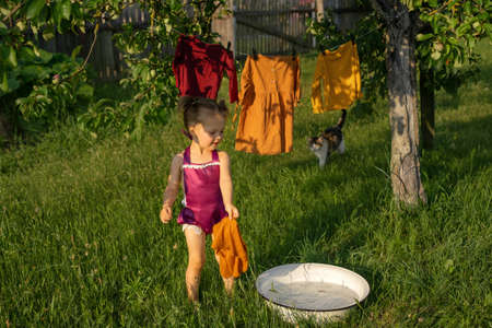 A cute girl splashes in a basin with soapy water, washes clothes and hangs clothes for drying in the garden. Washing and drying of colored clothes in the outdoors. The Little Helper and his catの写真素材
