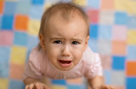 The baby cries standing next to his mother and looking up. A cute, tearful baby. Portrait of a little girl in tears on the background of a colorful blanket. fear in the child's eyesの写真素材
