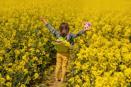 A girl in a rapeseed field with a backpack filled with modern educational popit and simple dimple toys. A bright yellow field of flowering canola. Sunny day. With open arms. stand with your backの写真素材