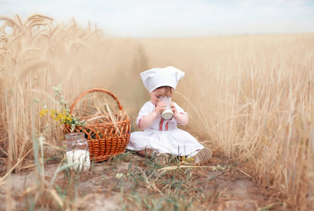 A healthy childhood in harmony with nature. happy little baby in folk clothes sits on a wheat field with a jar of fresh milkの写真素材