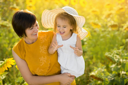 Emotional photo of a grandmother and granddaughter in a field of sunflowers. A cute little girl laughs and makes hand gestures. Love and understanding and a close connection between generations.の写真素材