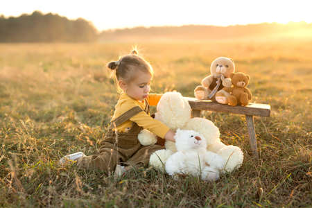 A little girl plays with teddy bears in a field at sunset. The child is lonely and his only friends are toy bears. Childhood in the country. Warm autumn evening with friendsの写真素材