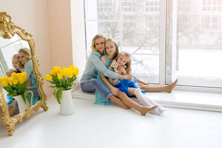 Mom hugs her daughters sitting at the window. A bouquet of yellow tulips in a vase stands by the window. Happy family. Flowers from daughters for mom on Mother's Day. child's day.の写真素材