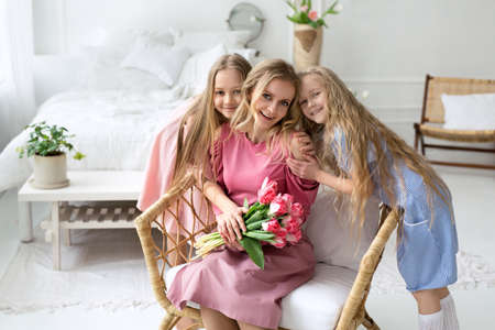 A mother with two daughters is sitting in a chair in the bedroom with a bouquet of pink tulips. Mother's love and tenderness. Mother's Day. Bouquet for your beloved mother.の写真素材