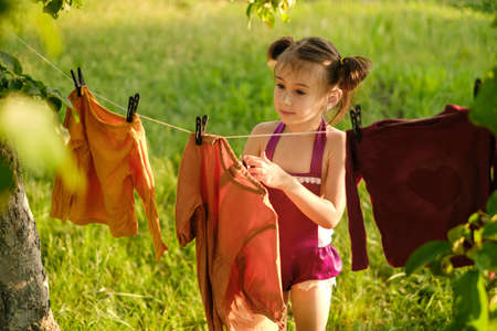 A cute little girl hangs a laundered dress to dry on a clothesline in the garden under a tree. Washing colored clothes with children's hypoallergenic safe washing powder. Rinses for baby clothesの写真素材