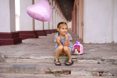 A girl with a pink heart-shaped balloon is sitting on the city steps with a toy dog in a portable basket. The child received his birthday gift.の写真素材