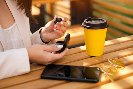 A woman takes out wireless bluetooth headphones from a charging case. A smartphone, sunglasses and a yellow cup of coffee are on the table. Time to listen to music and relax. earphones are modernの写真素材