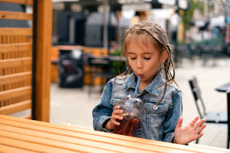 A little girl drinks fresh sweet lemonade from a straw while sitting at a table in the open air in a city cafe. A refreshing children's summer drink. Funny girl with pigtailsの写真素材