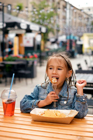 A little stylish girl in a denim jacket eats harmful fast food outdoors on the terrace of a cafe. The child is happy and enjoys eating fried chicken, washed down with a refreshing summer lemonadeの写真素材