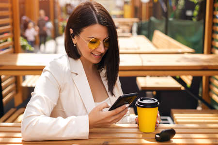 A young beautiful woman is sitting at a table in a cafe on the terrace, drinking her coffee, writing messages in her smartphone and listening to music through wireless headphones. A business womanの写真素材
