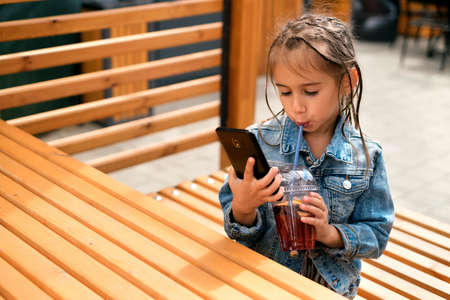 A little girl drinks a sweet summer drink lemonade through a straw and looks at her mobile phone. A child goes through online training on the phone while sitting in a city cafe in the open airの写真素材