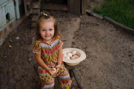 A little girl is sitting on a chair near the chicken coop and holding a chicken egg in her hands, smiling sweetly. A hat filled with eggs next to a child on a benchの写真素材