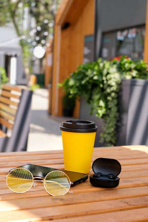 a glass of coffee with modern wireless headphones and a smartphone are lying on a table in an outdoor cafe. Work in the open air. Free time for recreationの写真素材