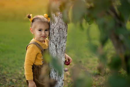 A little girl hugs a tree trunk on a warm autumn evening at sunset, holding a ripe red apple in her hand. A girl in a jumpsuit with bows in her hair. Life in the village.の写真素材