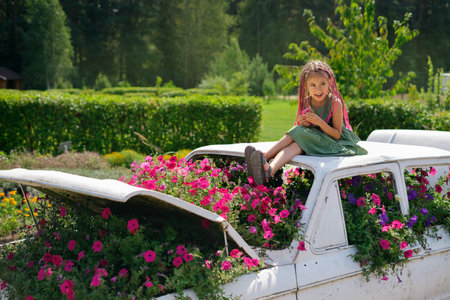 A girl with African pink pigtails from kanekalon is sitting on a designer landscape car with flowers. A flower garden of petunias in the garden. Funny kid. A flowerbed with petunia flowers. zizi pigtaの写真素材