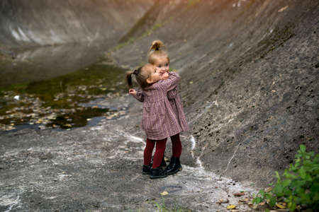 Two cute girls are hugging on the rocks in a rocky area. Girls like to play together. The children are dressed in identical dresses. They are as close as sisters.の写真素材