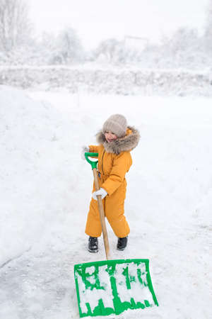 Girl in jumpsuit cleans snow big shovel. Snow removal after heavy snowfall. The six-year-old kid is happy to clean up the snow driftsの写真素材
