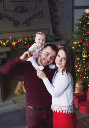 A beautiful young family with a small child is standing by the Christmas tree and fireplace. A little girl is sitting on her father's shoulders, and her mother is standing next to herの写真素材
